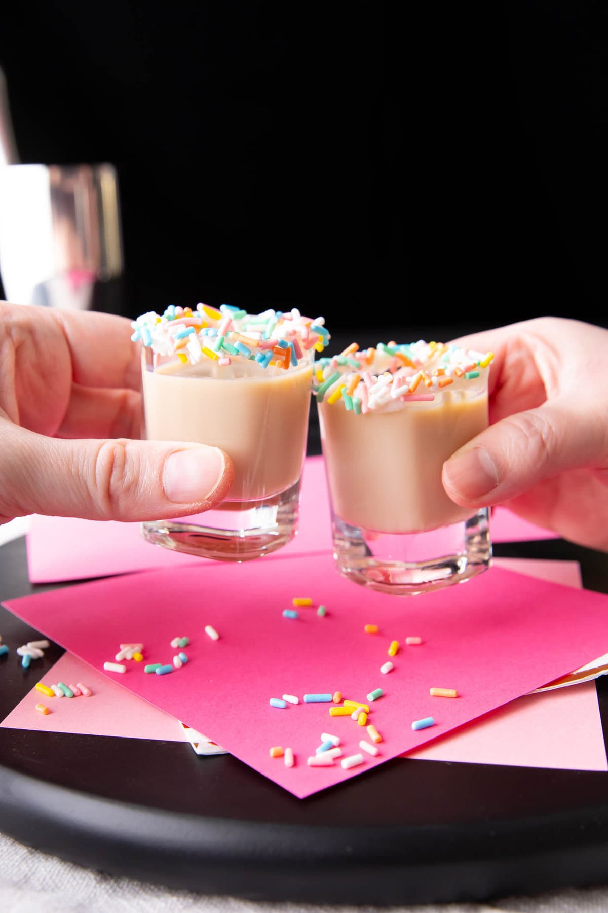 Two hands holding birthday cake shots, clinking shot glasses