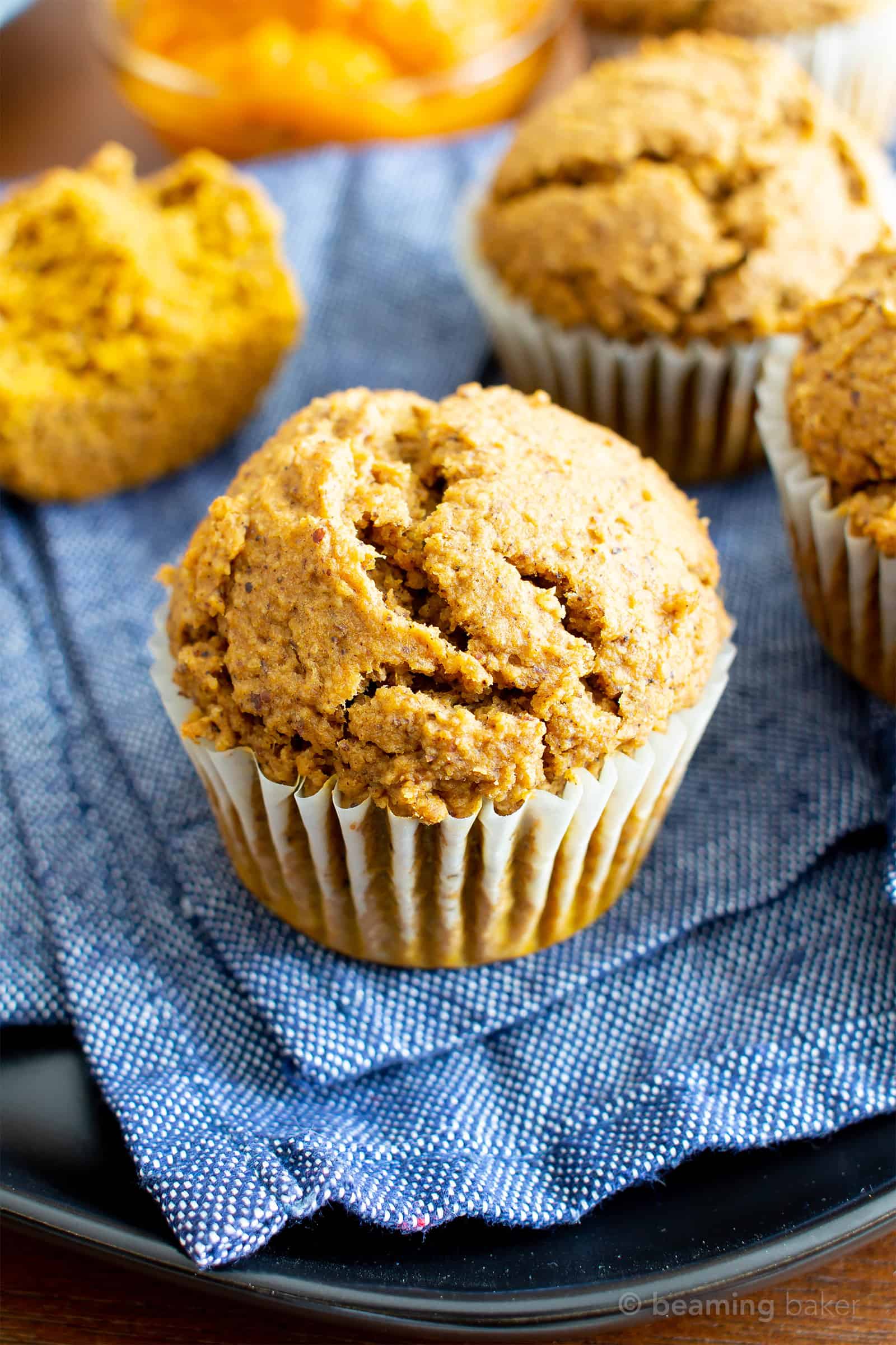 Several vegan gluten free pumpkin muffins on a blue cloth with a bowl of pumpkin puree and a split muffin in the background