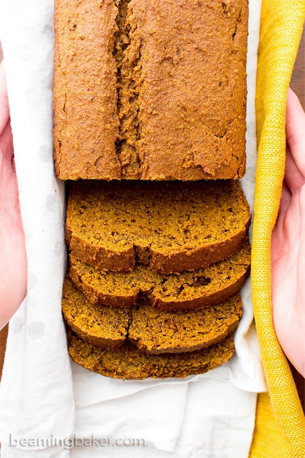 Overhead picture of vegan gluten free pumpkin bread loaf with several slices in the front and two hands holding in a white and yellow cloth